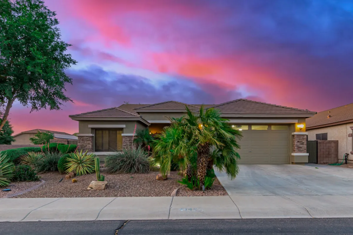 The image showcases the home's inviting exterior, highlighted by elegant palm trees, a warm sunset sky, and a welcoming front porch. The desert landscaping and stone accents reflect the region's natural beauty and low-maintenance lifestyle.