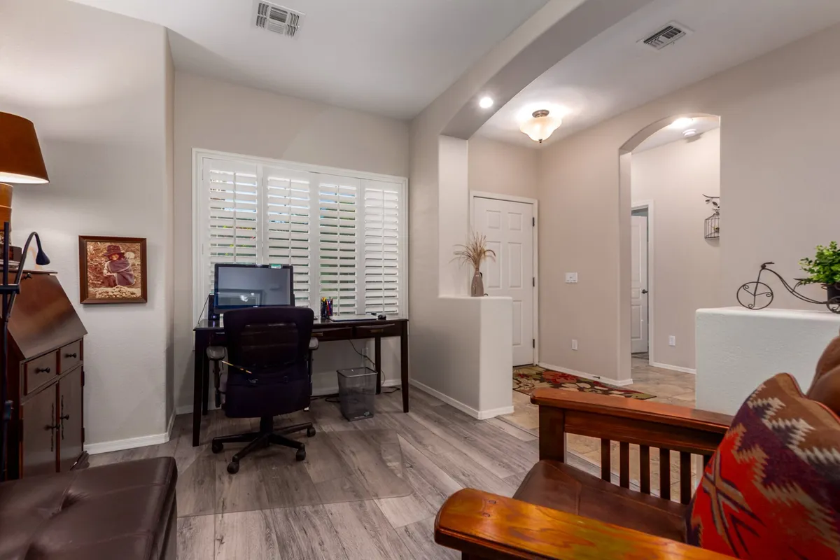 An interior den setup featuring a desk with a computer, a wooden chair, framed artwork, and a doorway to the hallway. The space is filled with natural light and designed for productive work sessions or video calls, as highlighted by the modern touches and cozy atmosphere.