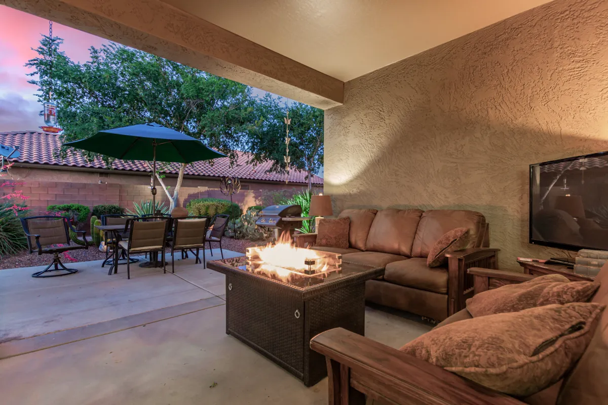 A welcoming backyard patio area featuring a comfortable seating arrangement around a fire table, lush green plants, an umbrella for shade, and a dining set perfect for outdoor meals. The warm, inviting ambiance is highlighted by beautiful sunset views.