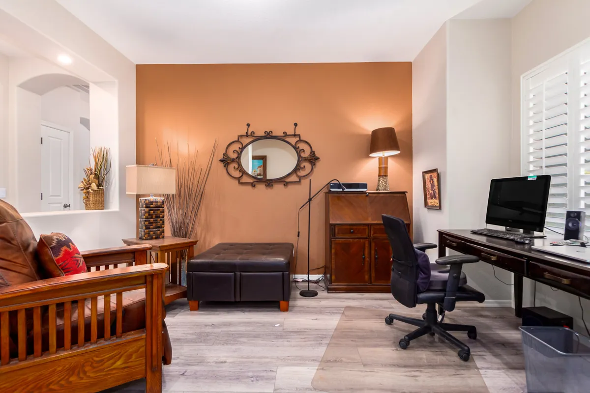 A well-lit den or work area featuring a wooden chair, decorative mirror, table lamp, and a desk with computer. The ambiance is comfortable and productive, perfect for reading, working from home, or focused activities. The styling is both modern and homey, aligning with the section’s emphasis on versatility.