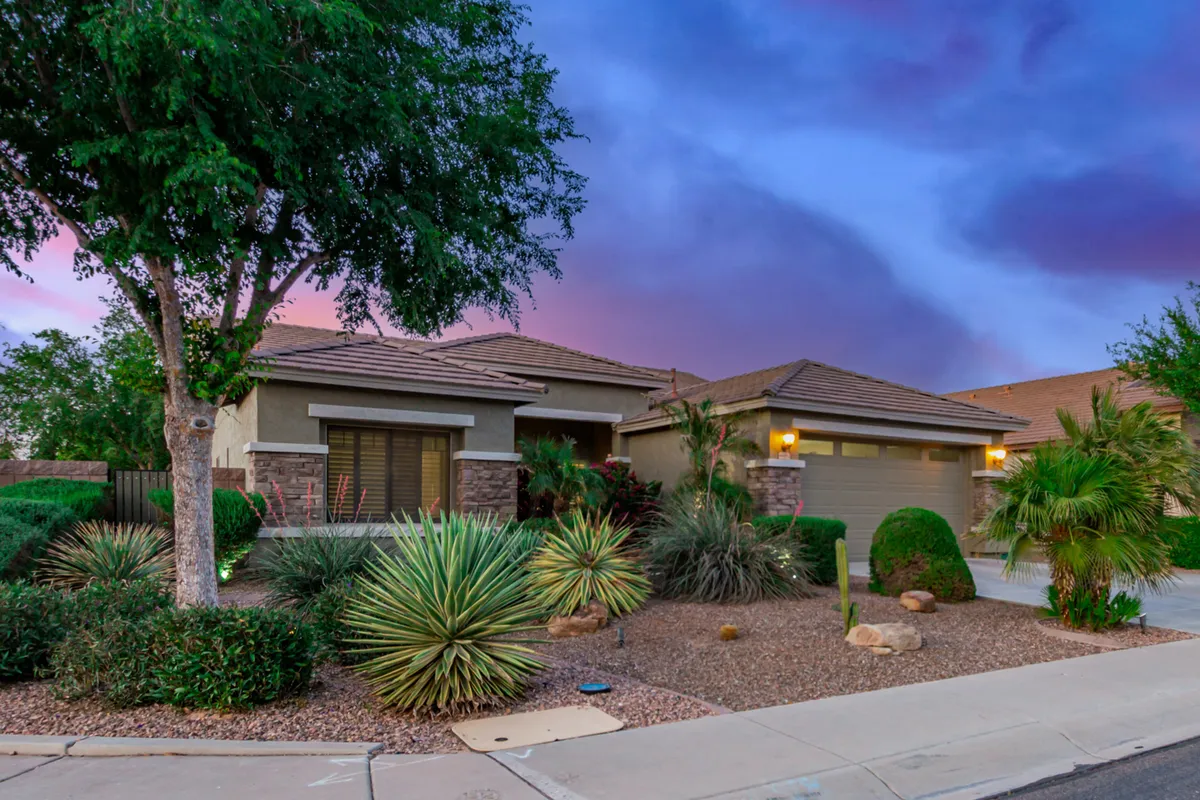 The front elevation of a home with well-maintained front yard landscaping, showcasing desert plants and a prominent garage door. The scene captures a relaxed, suburban atmosphere with the home bathed in warm sunset light.