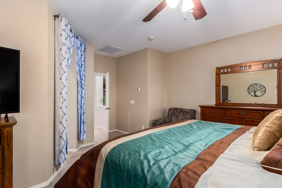 A spacious bedroom with teal and brown bedding, a wooden dresser with a mirror, a mounted TV, and a decorative curtain. The room is thoughtfully designed for relaxation and functionality, making it ideal for winding down after a long day.