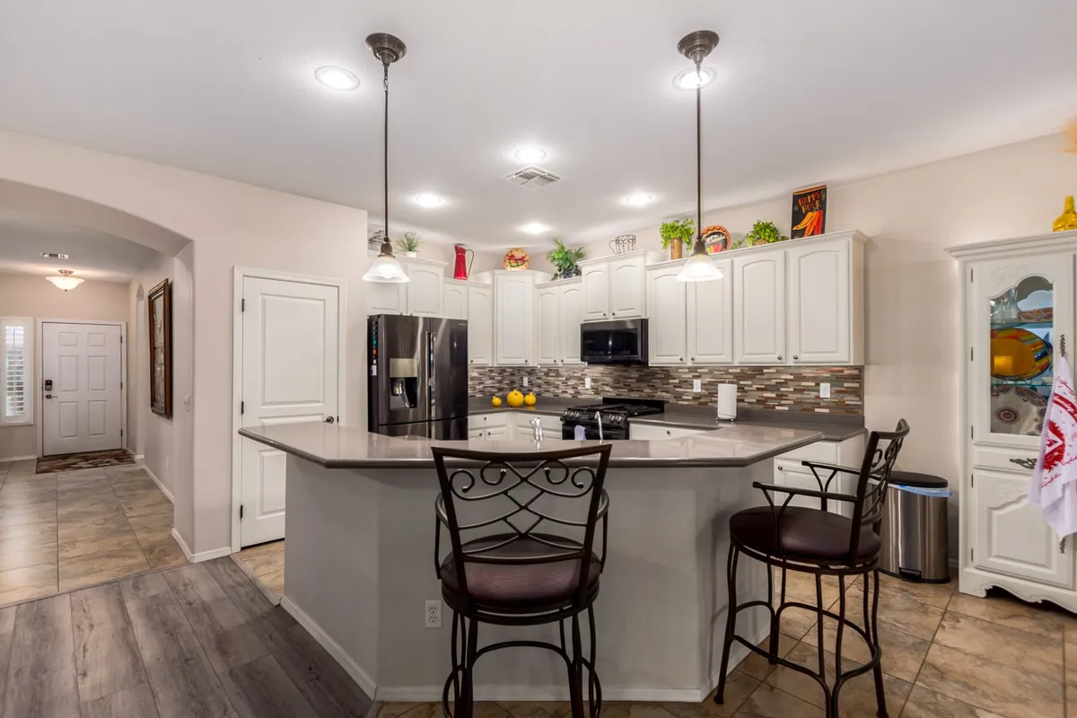 This image showcases the heart of the kitchen, highlighting the spacious island with bar stools, modern cabinetry crowned with molding, elegant mosaic tile backsplash, and high-end appliances. The arrangement suggests both functionality and a space designed for gathering and entertaining.