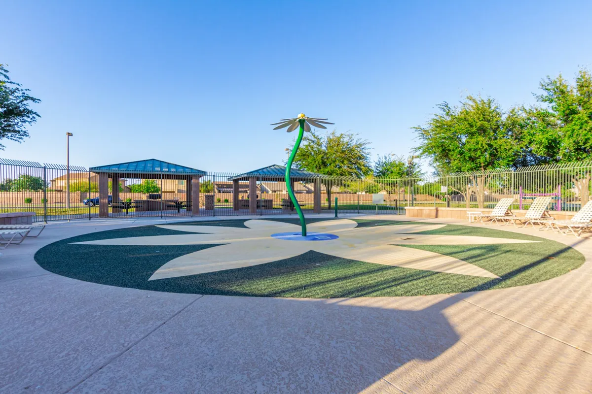 A playful splash pad with water features, surrounded by lounge chairs, shade structures, and mature trees. Children are seen playing in the water while families relax in the shaded seating, highlighting a social and active environment.