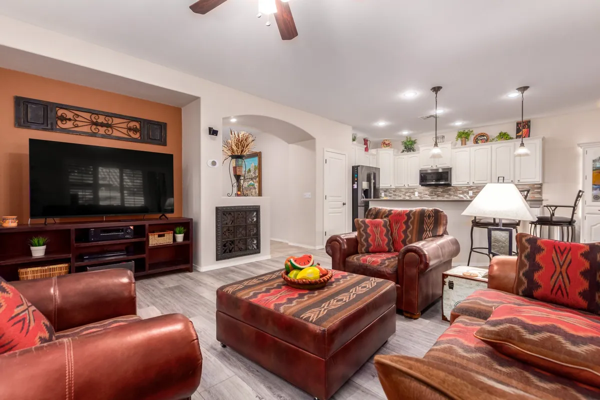 A sunlit great room featuring a large television, cozy seating area, colorful ottoman, and decorative wall art. Natural light floods the room, creating a warm and inviting space ideal for relaxing, watching movies, or entertaining guests. The arrangement and décor convey a sense of comfort and sociability.