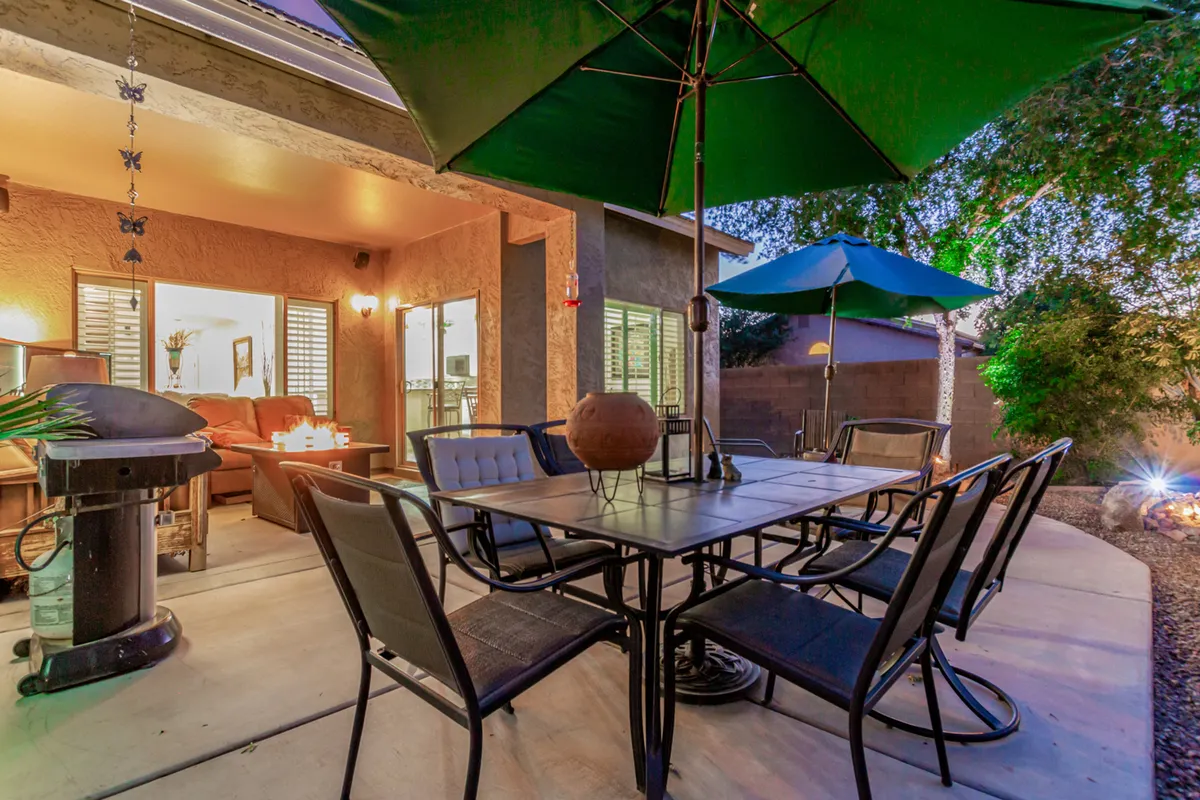 A welcoming exterior scene featuring an outdoor dining table with green umbrellas and a grill, set amidst a landscaped patio area. The space is clearly designed for social gatherings, sunset dinners, and starlit conversations, offering plenty of room for guests to relax and connect.