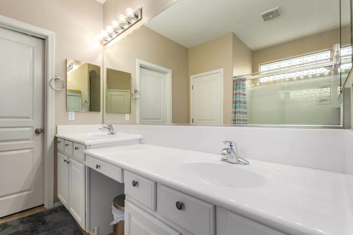 A bright bathroom featuring dual sinks with large mirrors, a walk-in shower area, and elegant light fixtures. The space is organized and calming, ideal for morning routines or unwinding in the evening.